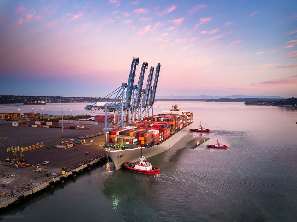 Large container ship guided by tugboats arriving at port terminal with tall gantry cranes at sunset