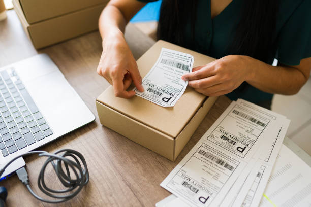 Person applying shipping label to cardboard box at desk with laptop and multiple package labels